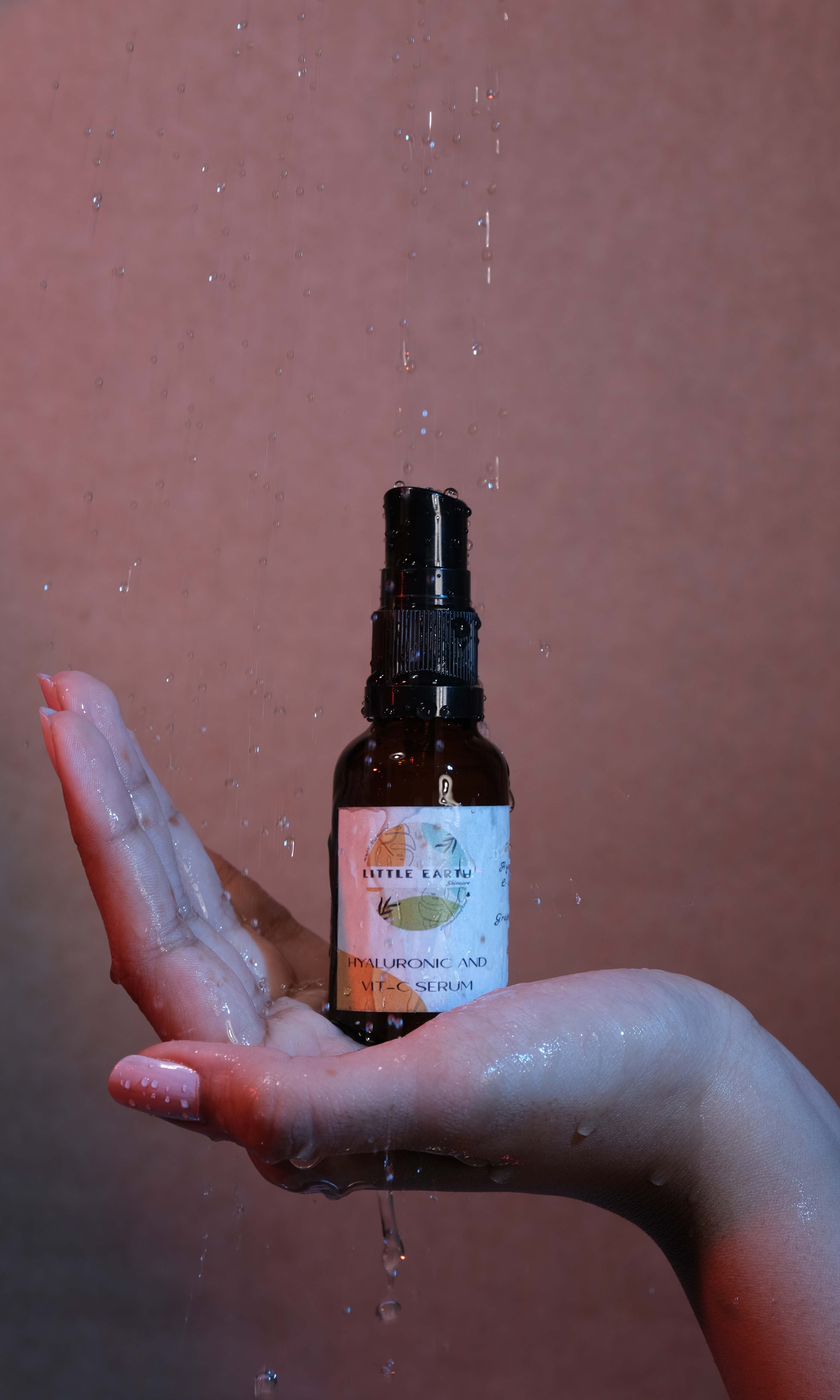 A bottle of Hyaluronic Acid and Vitamin C Serum held in a hand, with water droplets around indicating the serum is designed for skin hydration and brightening.